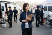 クリップボードを持った女性レーシングチームマネージャーが、レース会場のパドックで周囲を確認しながらスケジュール管理をしている様子。