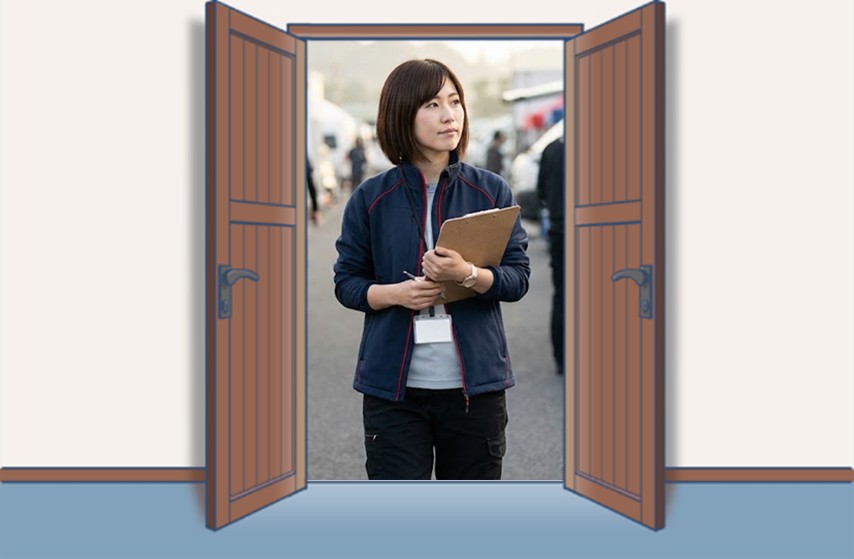 クリップボードを持った女性レーシングチームマネージャーが、レース会場のパドックで周囲を確認しながらスケジュール管理をしている様子。