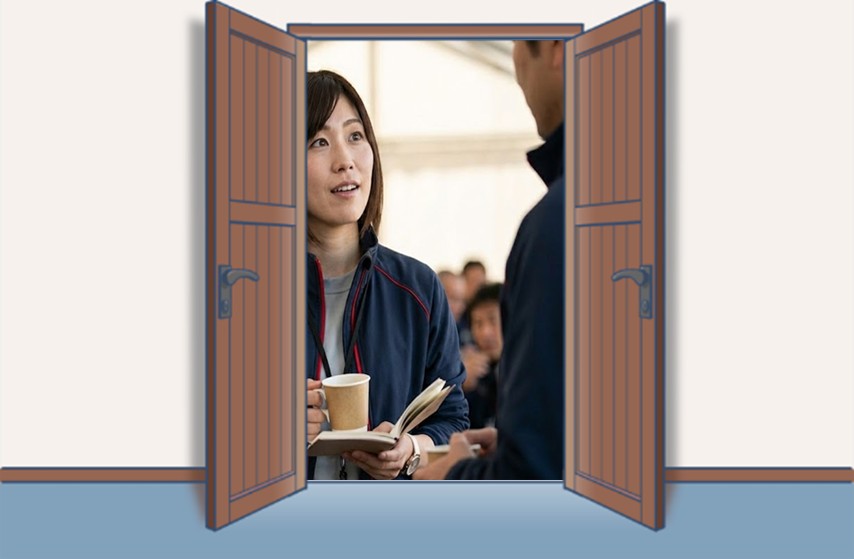 レーシングチームの女性マネージャーがパドック内のホスピタリティスペースで男性スタッフと話しながら、コーヒーとノートを手に打ち合わせをしている様子。